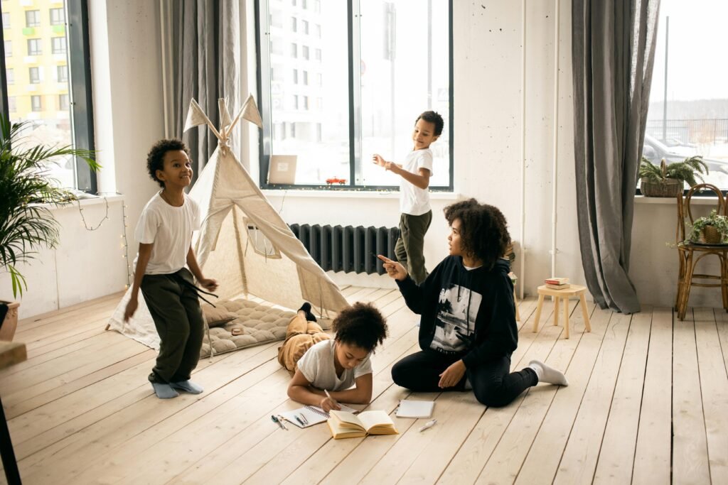 Full body of African American mother sitting on wooden floor near black kids playing in light room with blanket fort